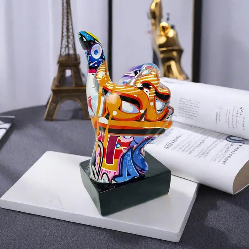 Thumbs-up hand gesture statue with colourful graffiti-style abstract patterns on a solid black square base.