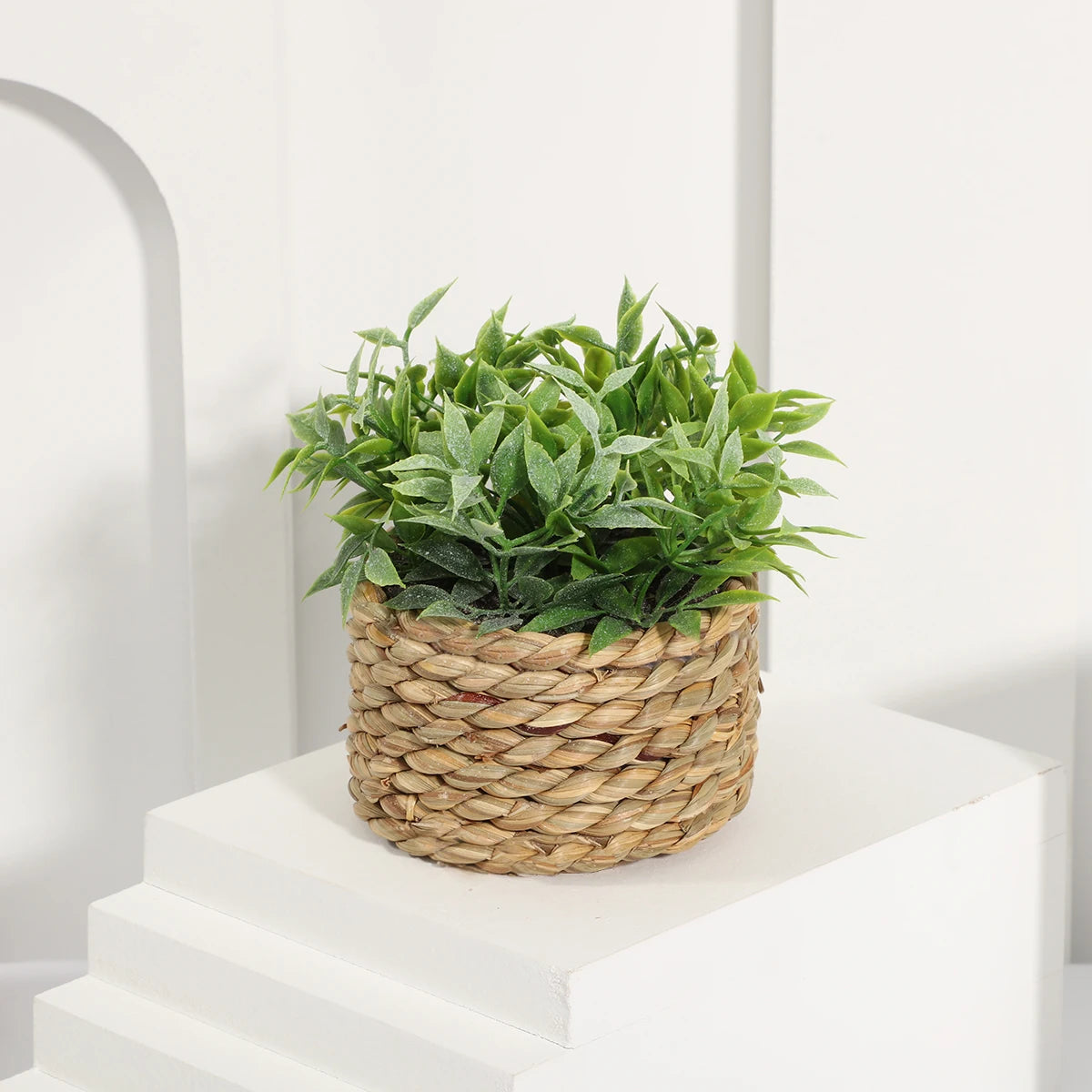 Small artificial green eucalyptus plant in a woven seagrass basket, sitting on a white tiered platform.
