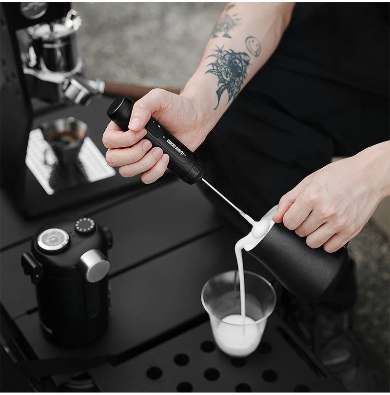 Handheld black electric milk frother being used to pour frothed milk from a matching black pitcher into a glass.