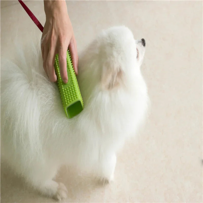 A hand uses a lime green silicone grooming brush to brush the white fur of a small Pomeranian-style dog.