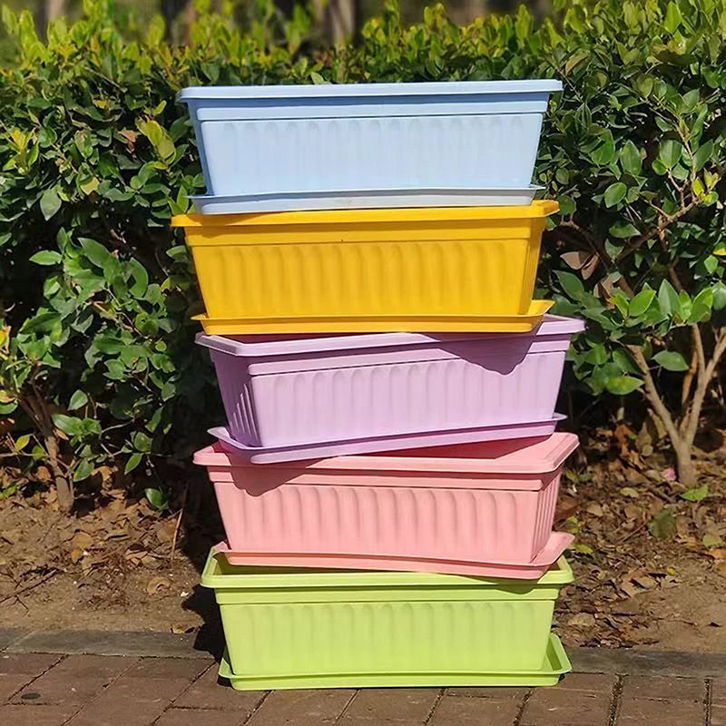 A stack of five rectangular plastic garden planters in blue, yellow, lavender, pink, and lime green on a brick path.