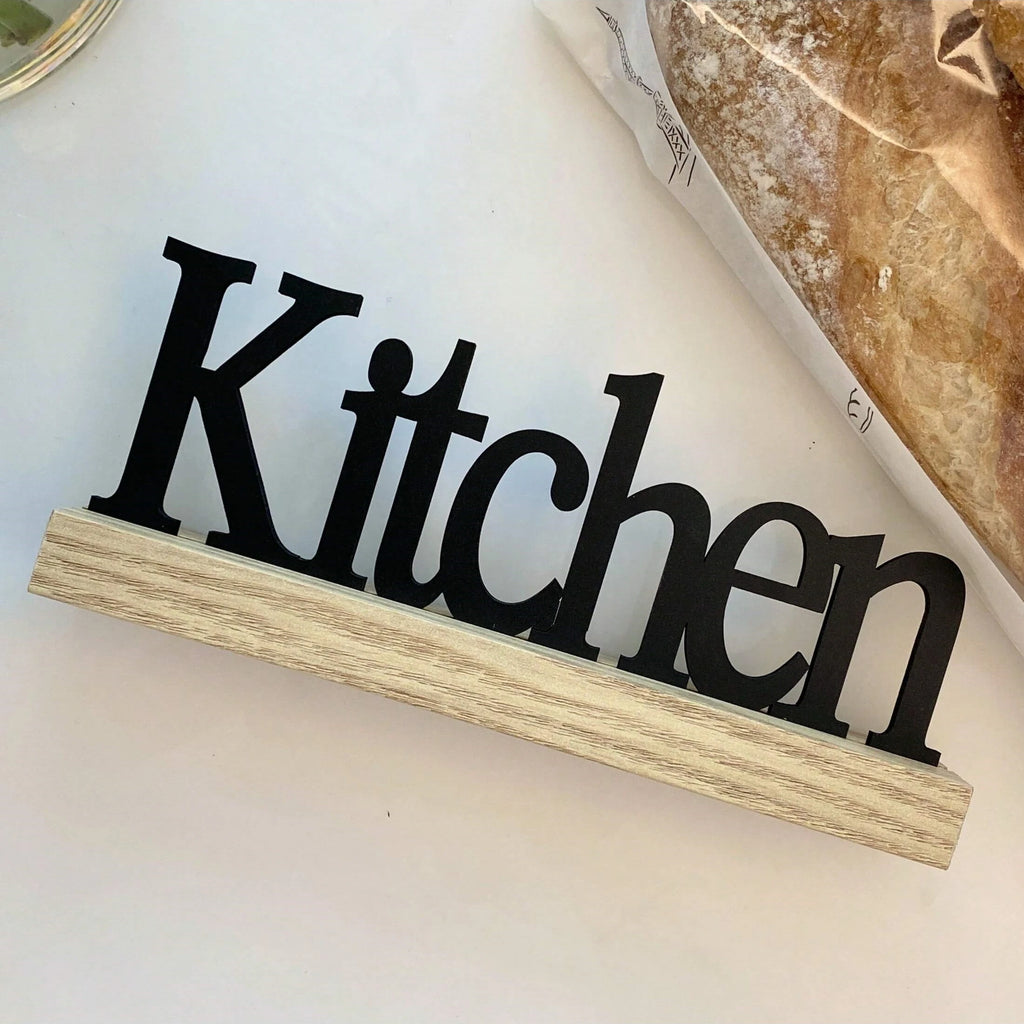 Black 'Kitchen' sign with elegant lettering standing on a light wood grain base.