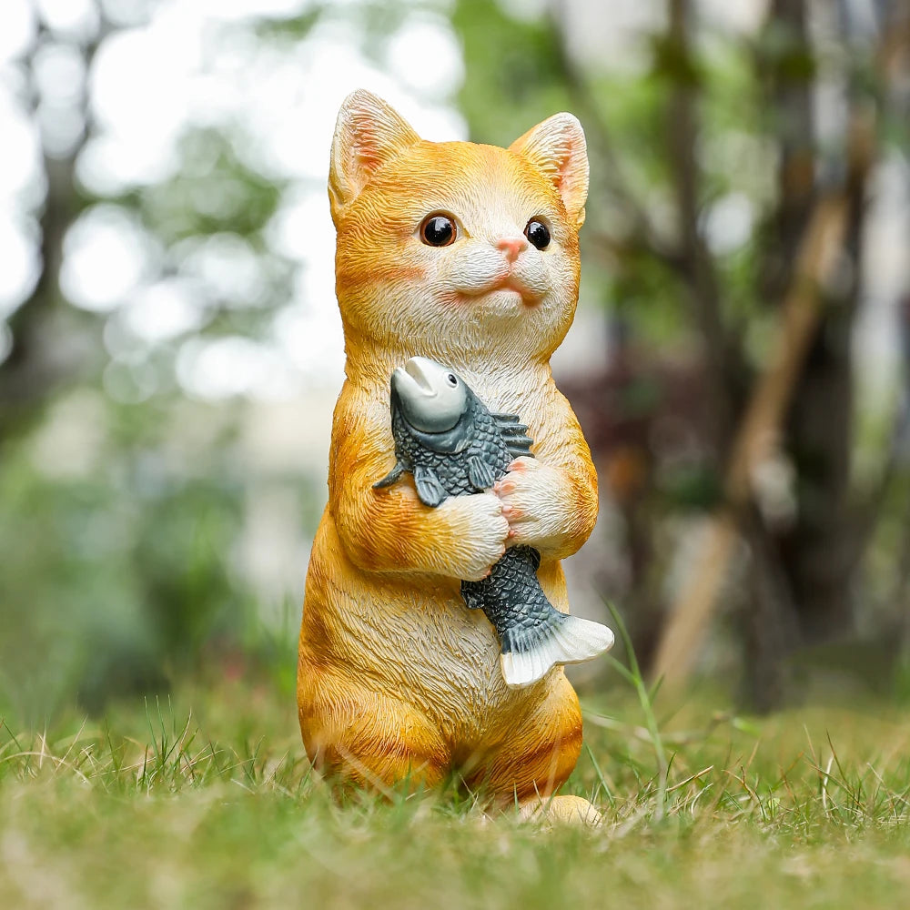 A charming, standing orange resin cat garden statue holding a small grey fish, set against a soft-focus garden backdrop.