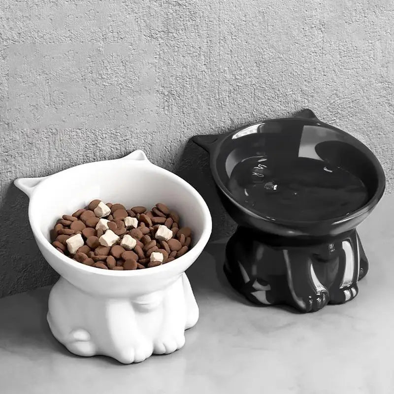 Two elevated, 15-degree tilted ceramic pet bowls in white and black, shaped like cute cats, one filled with dry food.