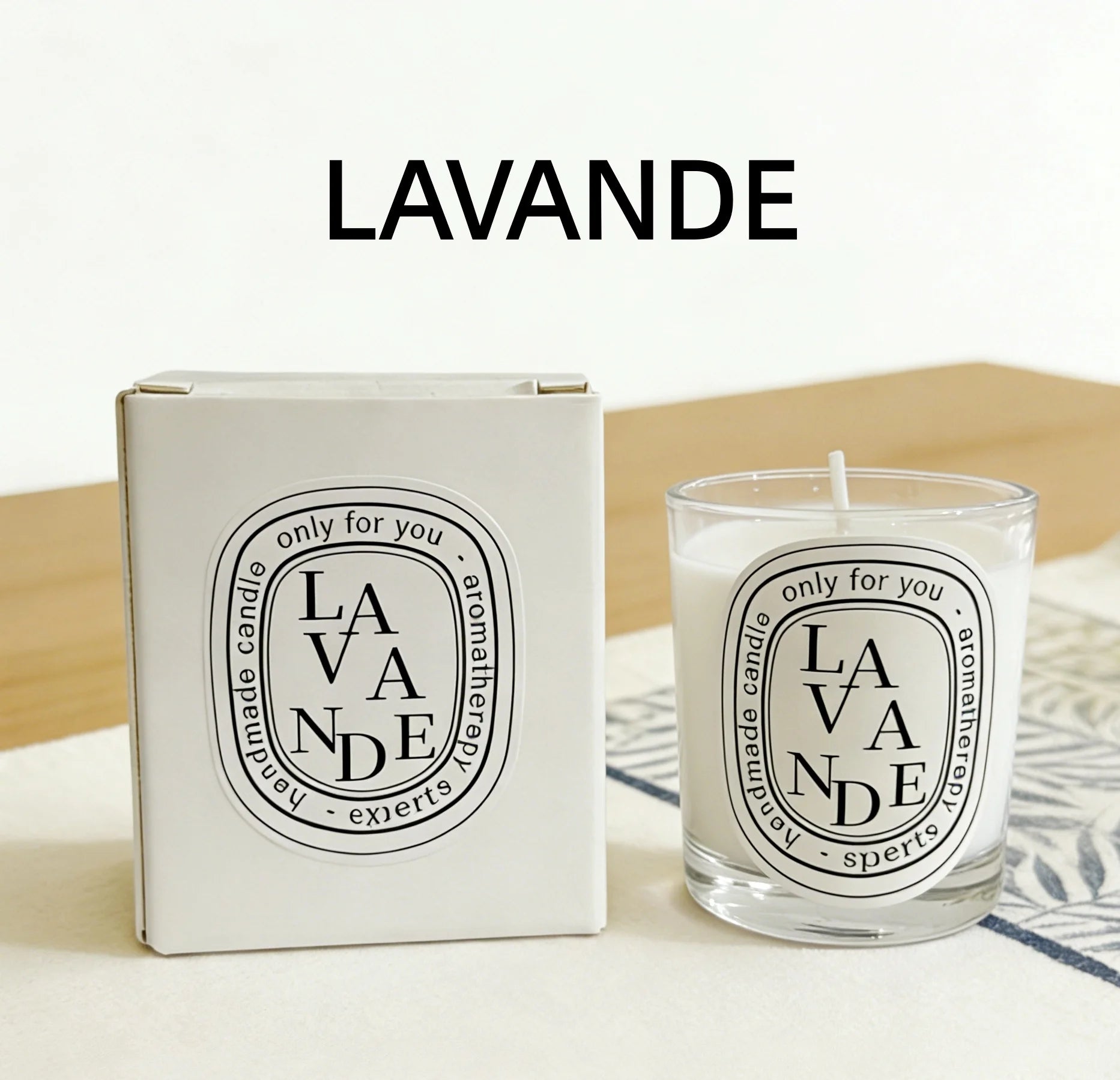 A white aromatherapy candle in a glass jar next to its matching white box, featuring a black oval "Lavande" label.