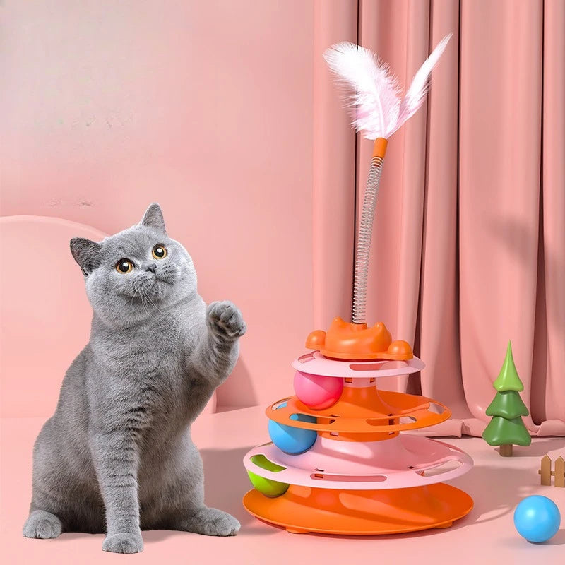 A grey cat plays with an orange and pink tiered track toy featuring rolling balls and a springy feather teaser wand.