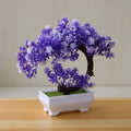 Artificial bonsai tree with purple flowers in a small, rectangular white pot on a wooden surface.