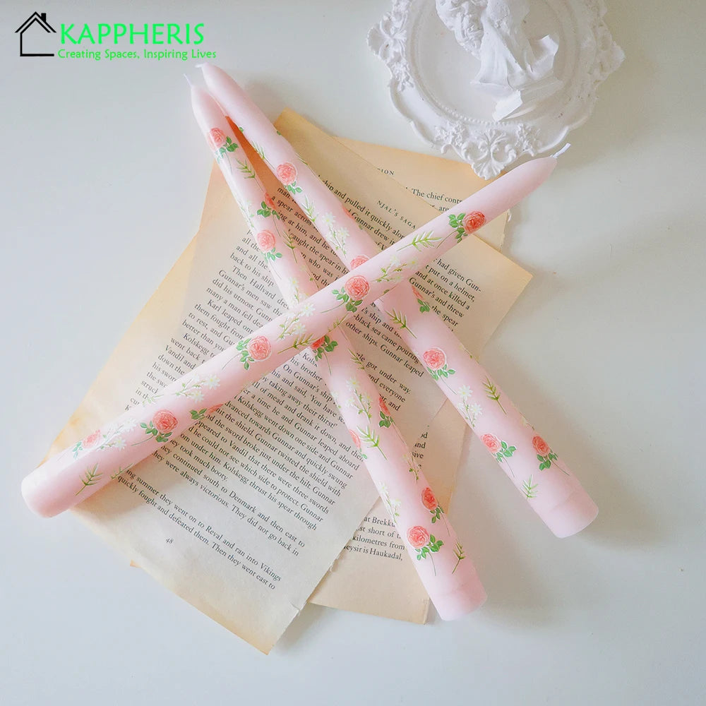 Three pale pink tapered dinner candles featuring a delicate floral rose pattern, laid across vintage book pages.