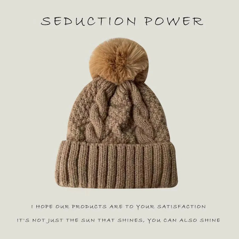 A tan cable-knit beanie hat featuring a fluffy faux fur pom pom and a ribbed turn-up cuff.