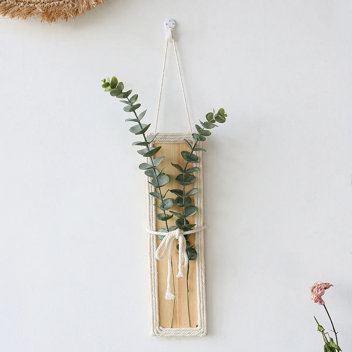 Bohemian wall hanging featuring a rectangular light wood panel edged in cream rope, holding two green eucalyptus stems.