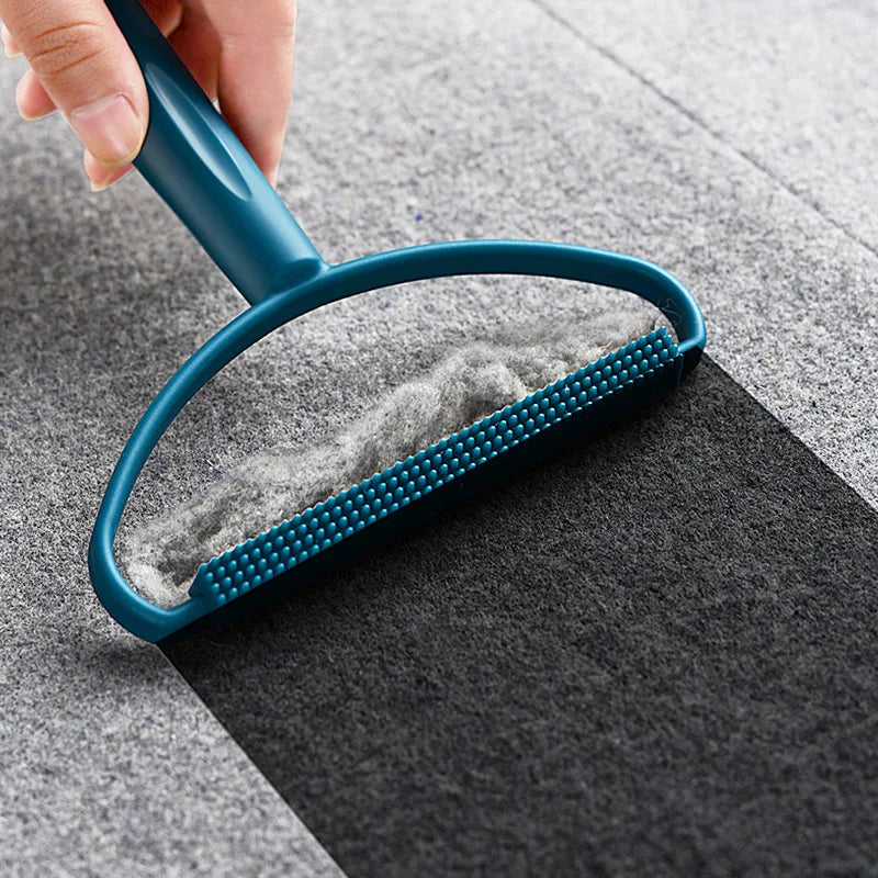 Hand holding a teal lint remover tool scraping grey fluff from a dark fabric surface.