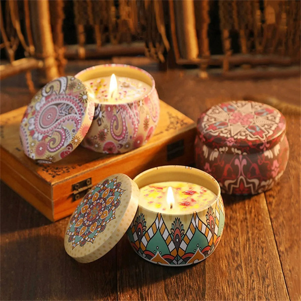 Two lit scented candles in decorative, colourful patterned metal tins with matching lids on a wooden surface.