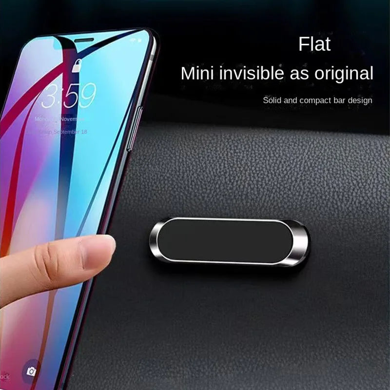Small, flat black and silver magnetic car phone mount attached to a textured dashboard next to a smartphone.