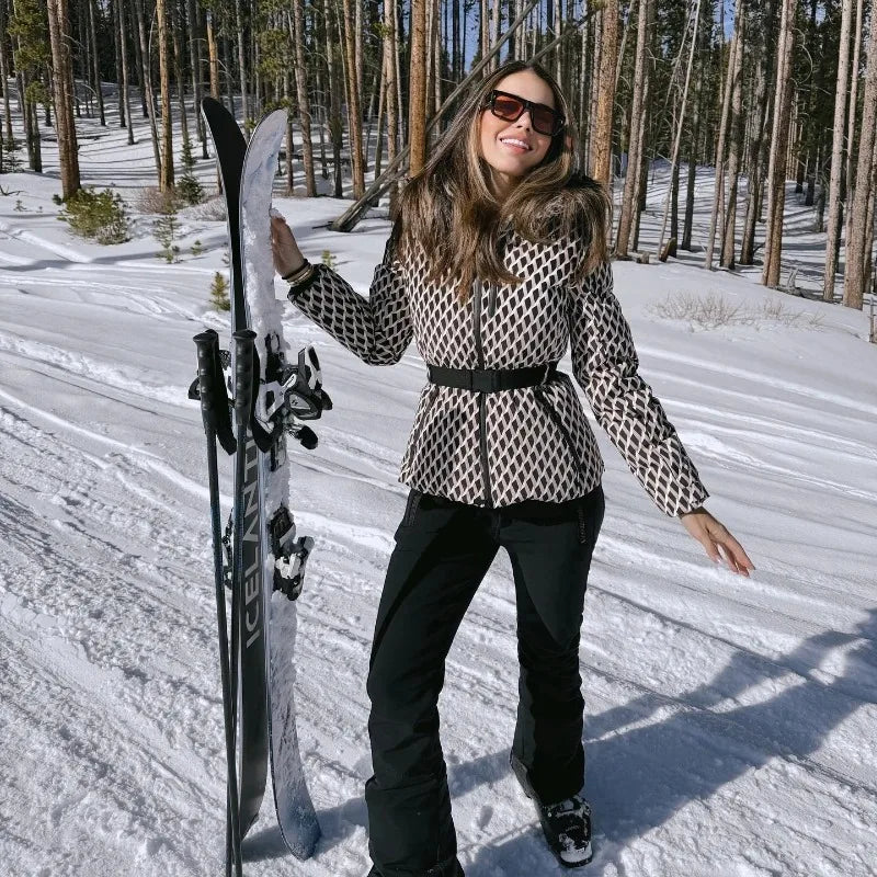 Woman in snowy forest wearing a patterned, belted, faux-fur-hooded ski jacket with black trousers and sunglasses.