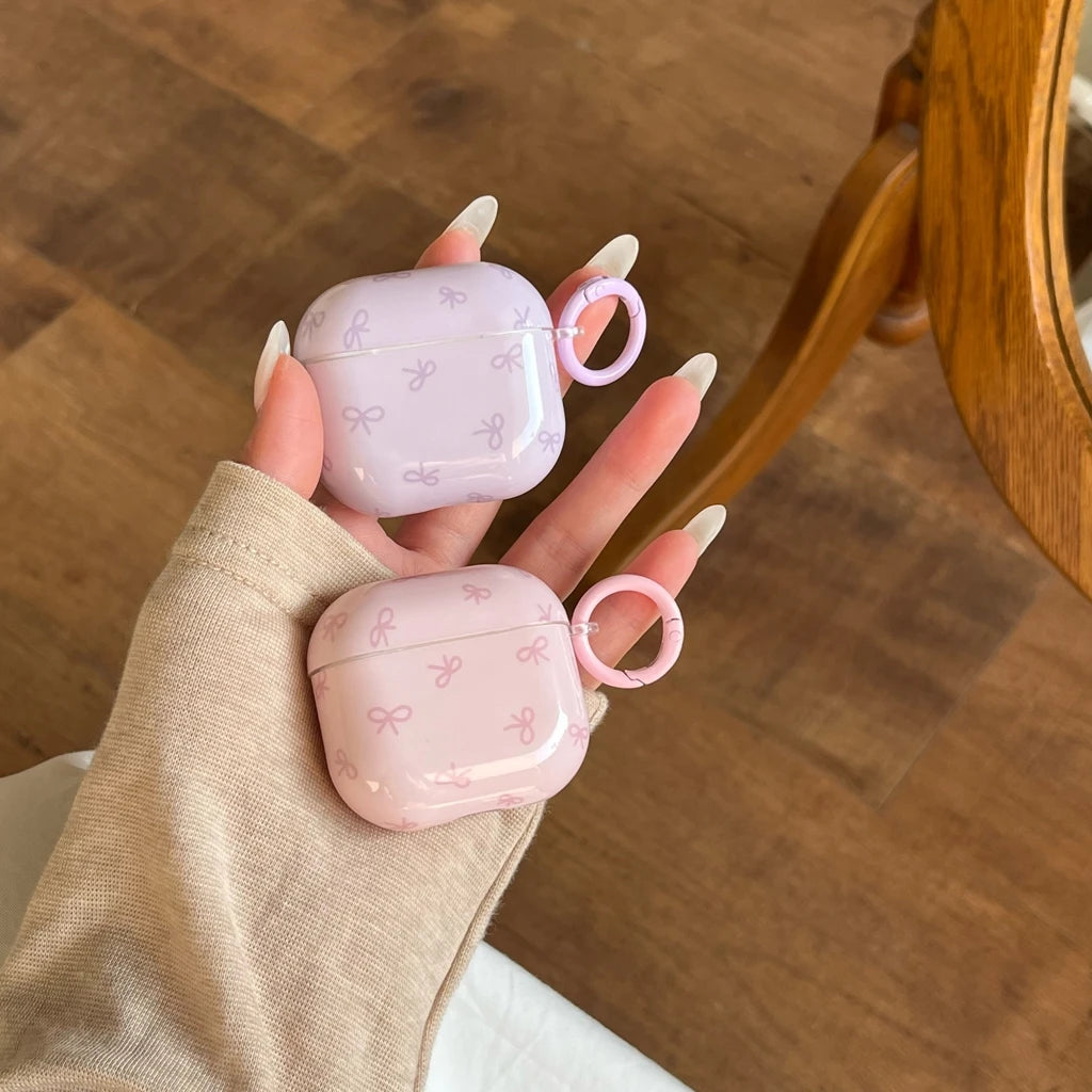Glossy pastel lilac and pink AirPods cases with a delicate bow print and integrated ring holder.