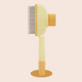 Pale yellow and orange pet grooming brush with fine metal teeth, standing upright on a matching round base.