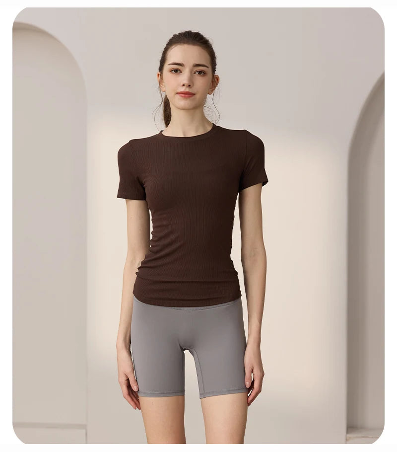 A woman wearing a dark brown ribbed short-sleeve fitted t-shirt and grey high-waisted activewear shorts.
