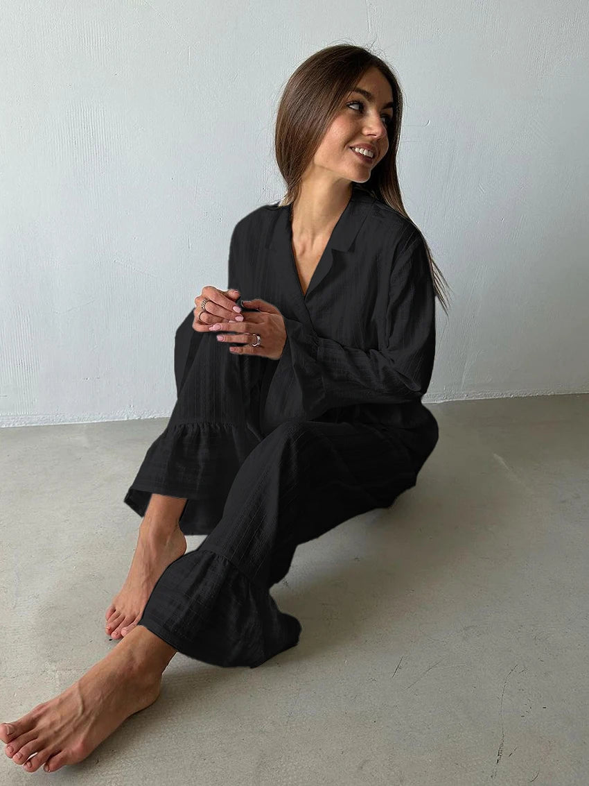 A woman wearing a black, long-sleeved lounge set featuring a collared shirt and wide-leg trousers with ruffled hems.