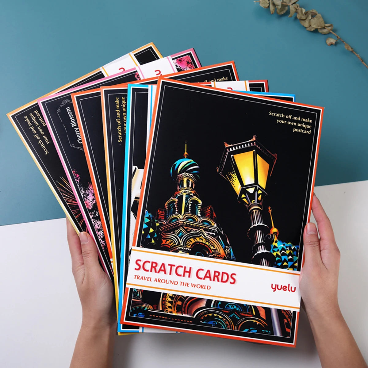 Hands holding a set of black scratch-off travel postcards featuring colourful, detailed world landmark designs.