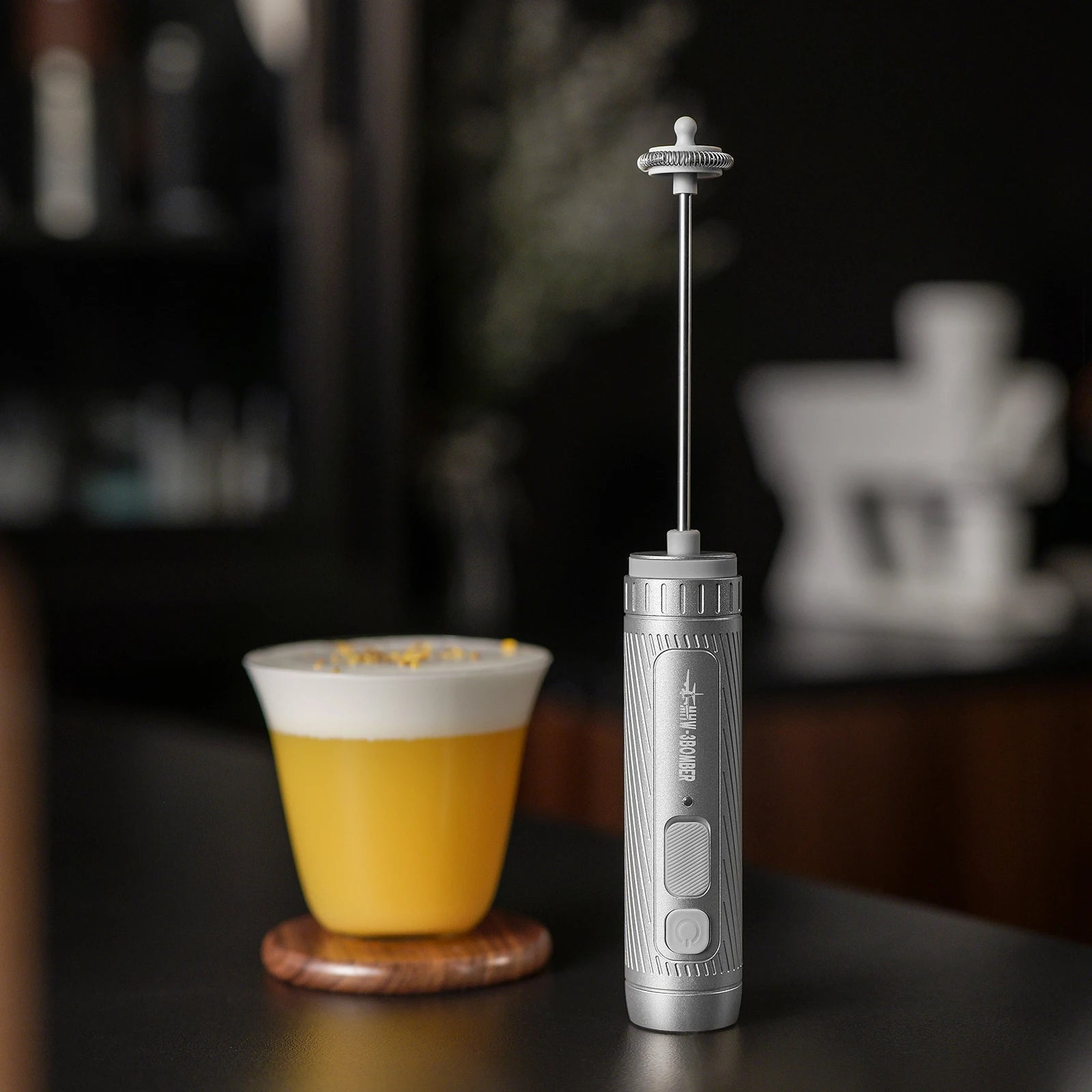 A handheld silver milk frother with a textured handle, standing next to a glass of yellow drink on a dark surface.