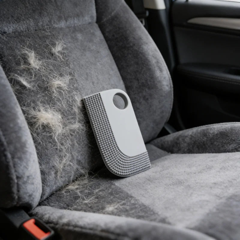 Grey rubber pet hair remover tool with a finger hole, placed on a fabric car seat partially covered in animal fur.