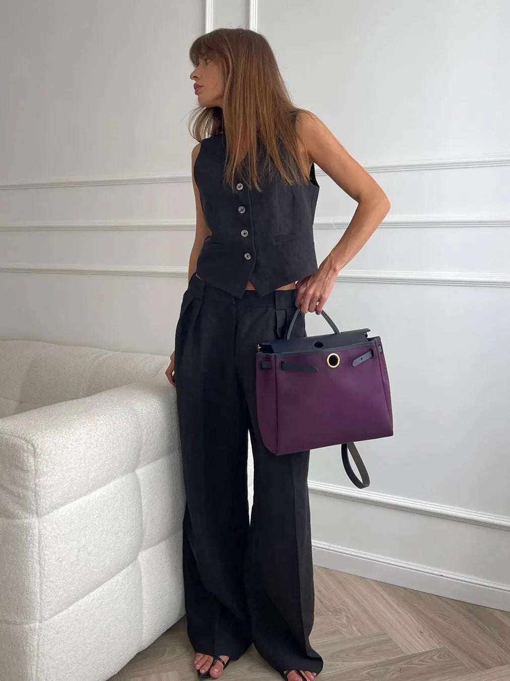 A stylish woman holds a structured, plum-purple handbag featuring a gold-toned circular clasp and dark top handle.