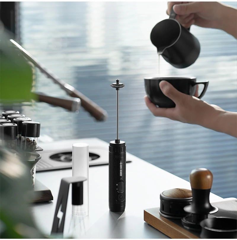 A handheld, matte black electric milk frother stands vertically on a modern kitchen counter with coffee accessories.