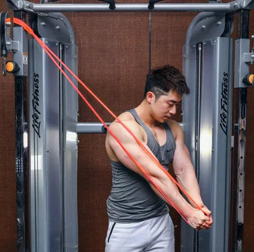 A man in a grey vest uses a long orange resistance band attached to a gym cable machine for chest exercises.
