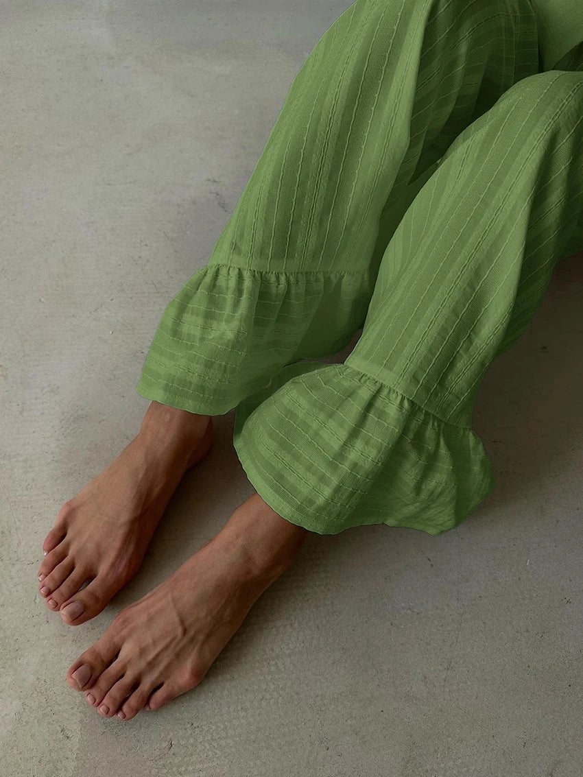 Sage green lounge trousers with subtle vertical stripes and ruffled hems, worn by a person seated on a grey floor.