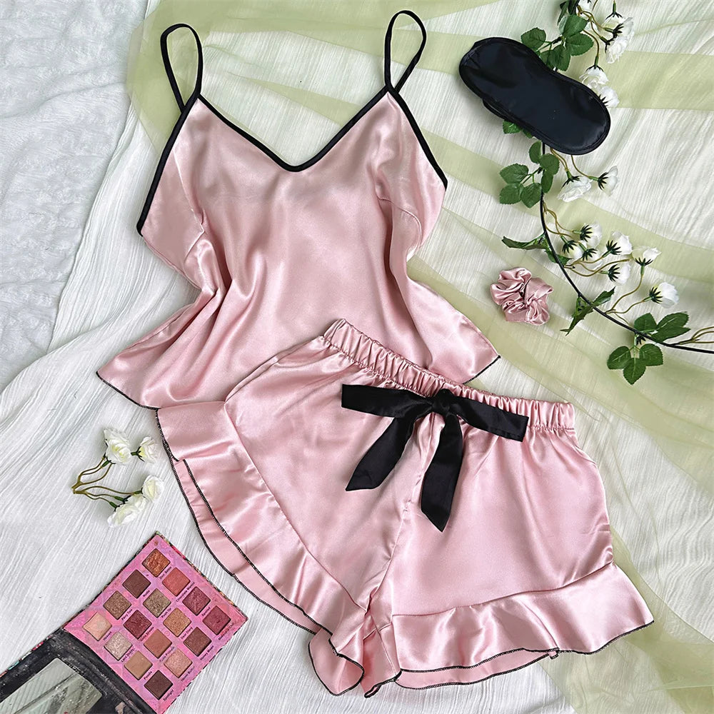 Pink satin pyjama set with camisole and ruffled shorts, featuring black trim, bow detail, eye mask, and hair scrunchie.
