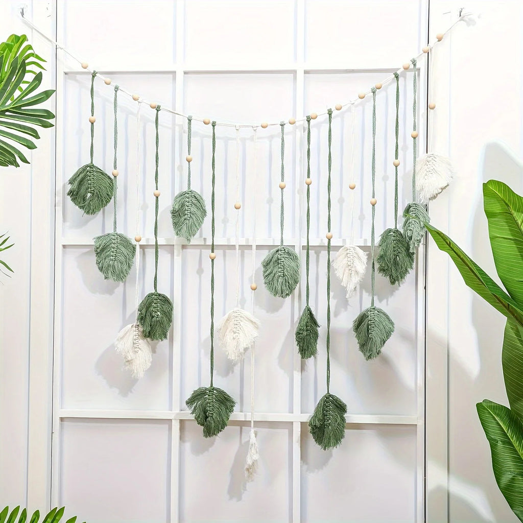 Boho macrame wall hanging featuring sage green and cream leaf motifs on strings decorated with wooden beads.