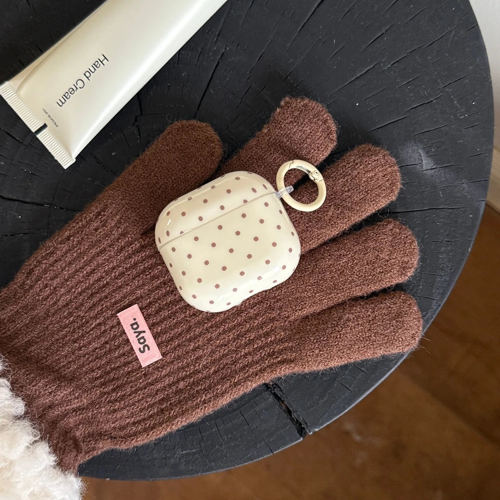 Cream-coloured polka dot AirPods case with keychain ring, resting on a brown knitted glove.