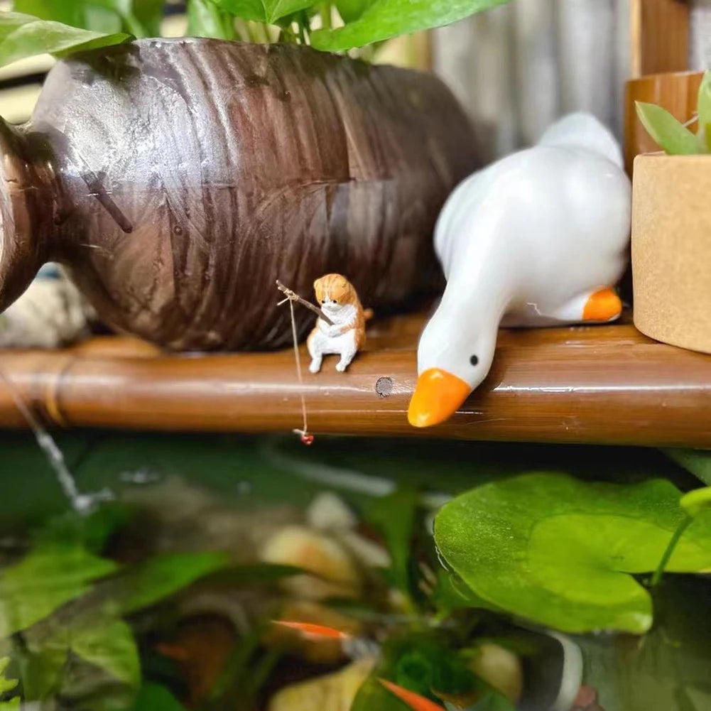 Tiny ginger tabby cat ornament fishing with a rod, sitting on a bamboo shelf next to a ceramic white goose.
