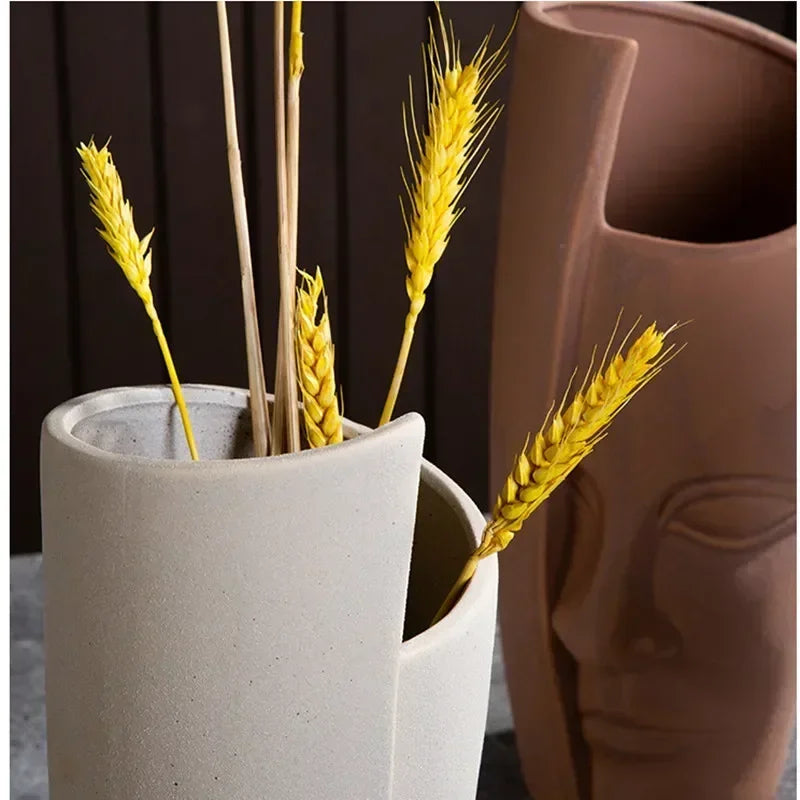 Two modern, sculptural face-shaped vases in grey and terracotta, featuring dried yellow wheat stems.