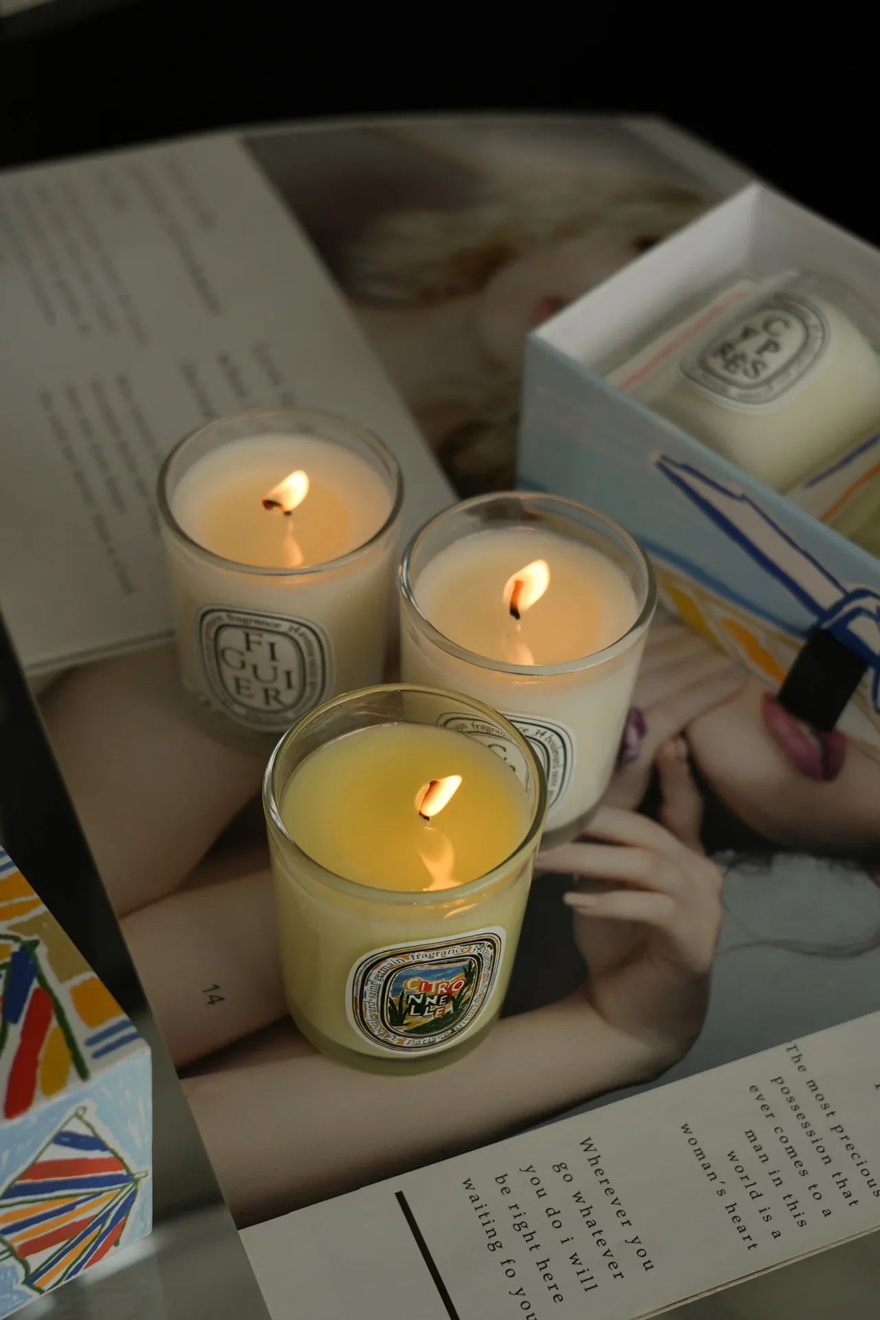 Three lit scented candles in glass jars with decorative labels, displayed on an open magazine.