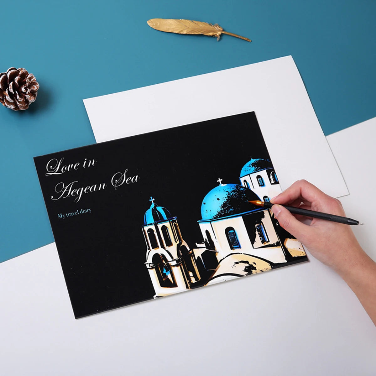 Hand using a stylus to reveal a colourful Greek island scene on a black "Love in Aegean Sea" scratch art travel diary.