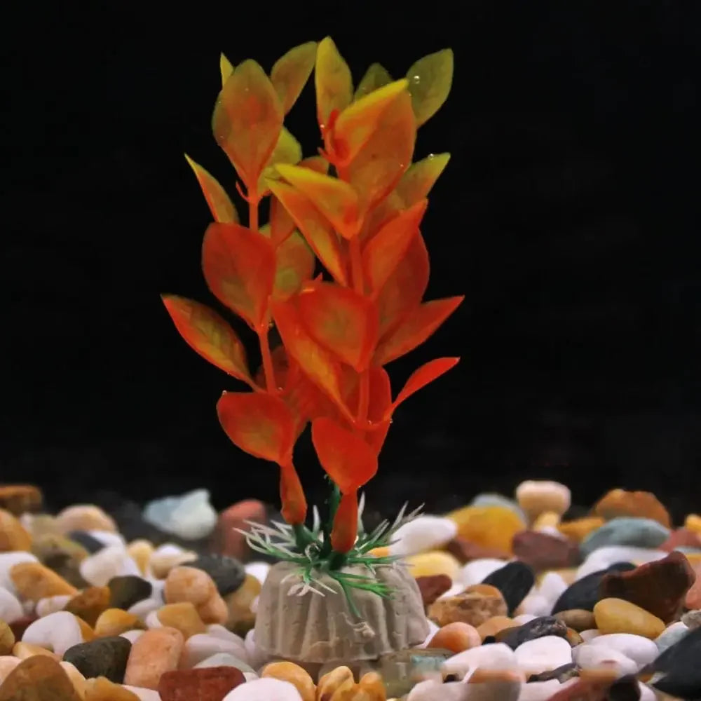 Artificial aquarium plant with orange and yellow leaves, set in a weighted stone-colored base on colorful gravel.