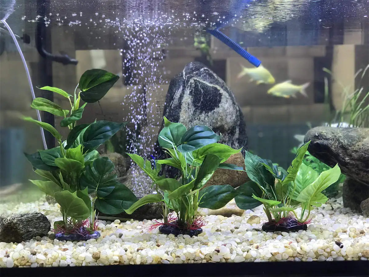 Three artificial green aquarium plants with broad leaves are arranged in a fish tank with light gravel and a rock.