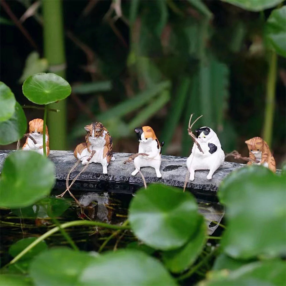 Five miniature, colourful cat figurines sitting on a stone edge, each holding a small fishing rod above a pond.