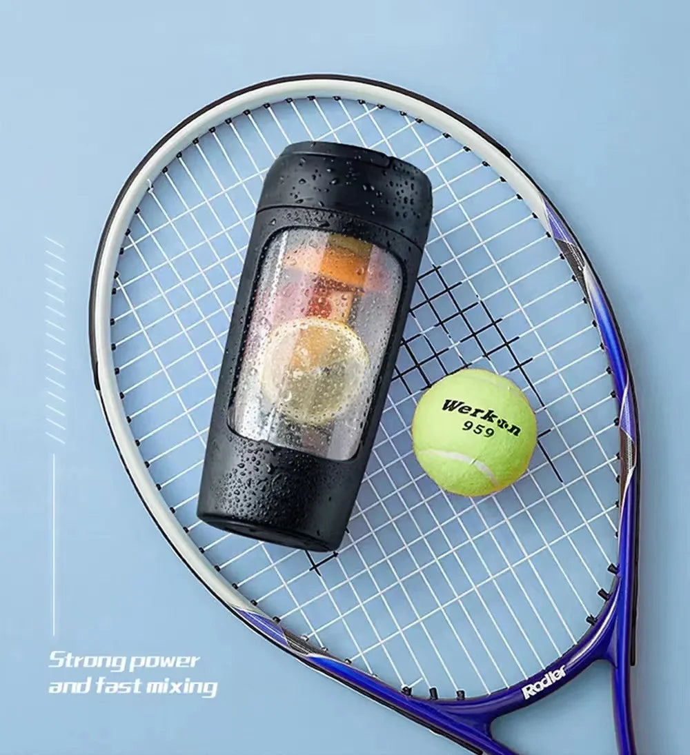 A black portable blender with a clear window containing fruit, resting on a tennis racket with a yellow tennis ball.