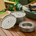 A set of titanium camping cookware on a wooden table, featuring a pot, steamer insert, and nested bowls.