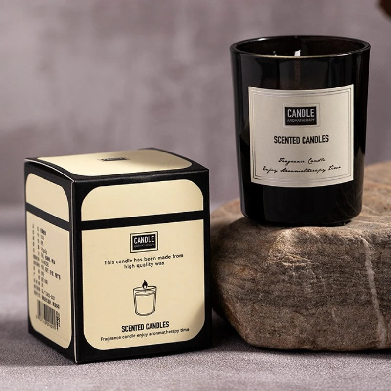 Black scented candle in a glass jar, paired with its elegant cream and black presentation box on a stone surface.