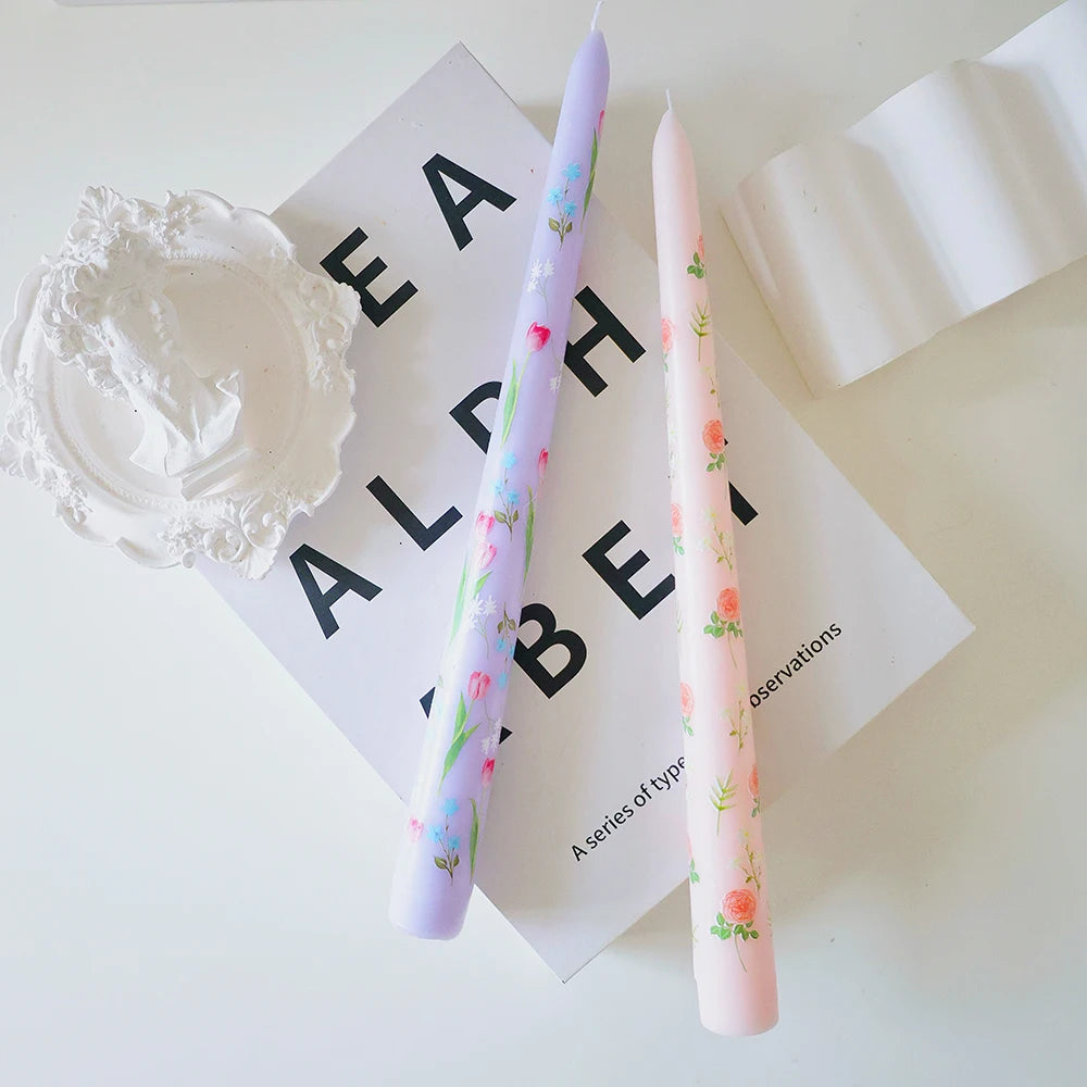 Two floral tapered dinner candles in pastel lavender and pink, displayed on a decorative book and white surface.