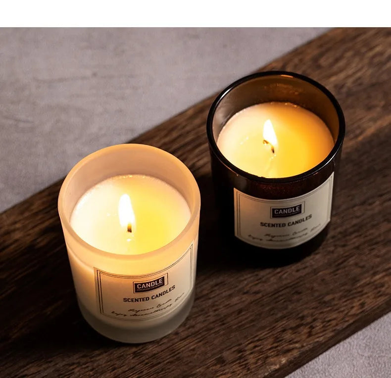 Two scented candles in frosted and dark glass jars, glowing with warm flames on a rustic wooden board.