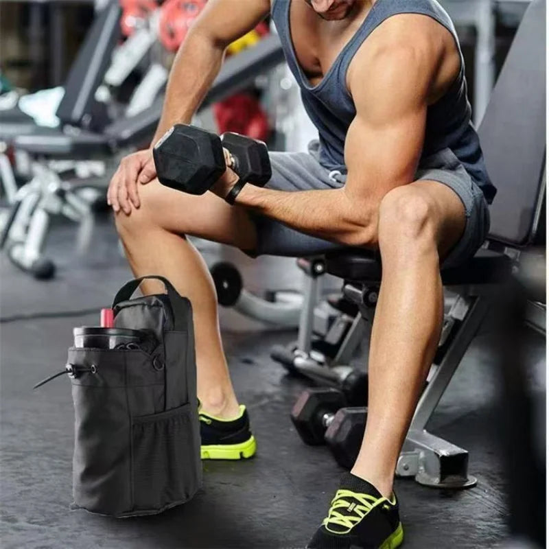 A black insulated gym water bottle cooler bag standing on a floor next to a man lifting dumbbells in a gym.