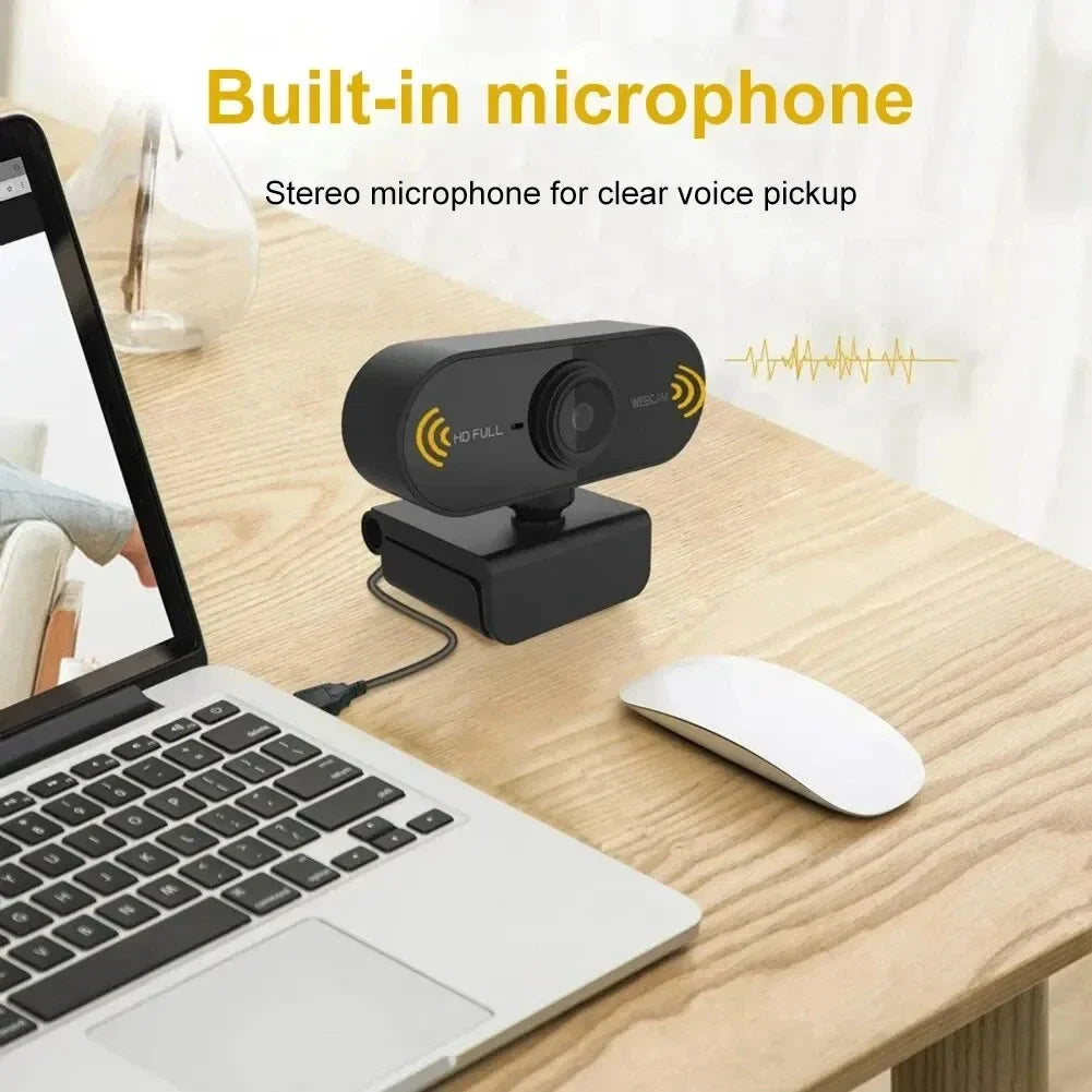 Black 1080p HD USB webcam with built-in microphone, sitting on a wooden desk next to a laptop and wireless mouse.