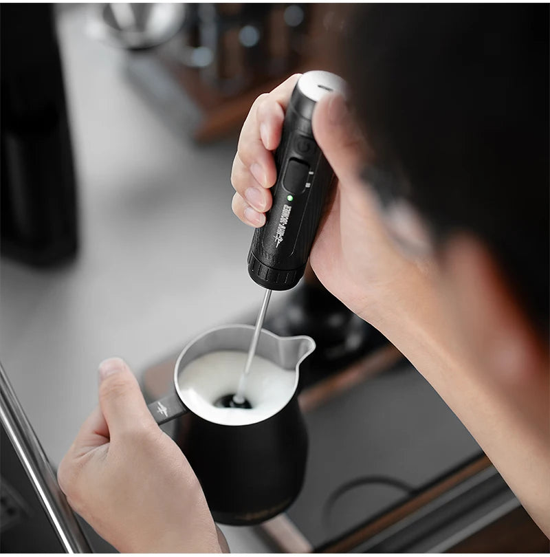 Black handheld electric milk frother in use with a matching black metal pitcher to foam milk for coffee.
