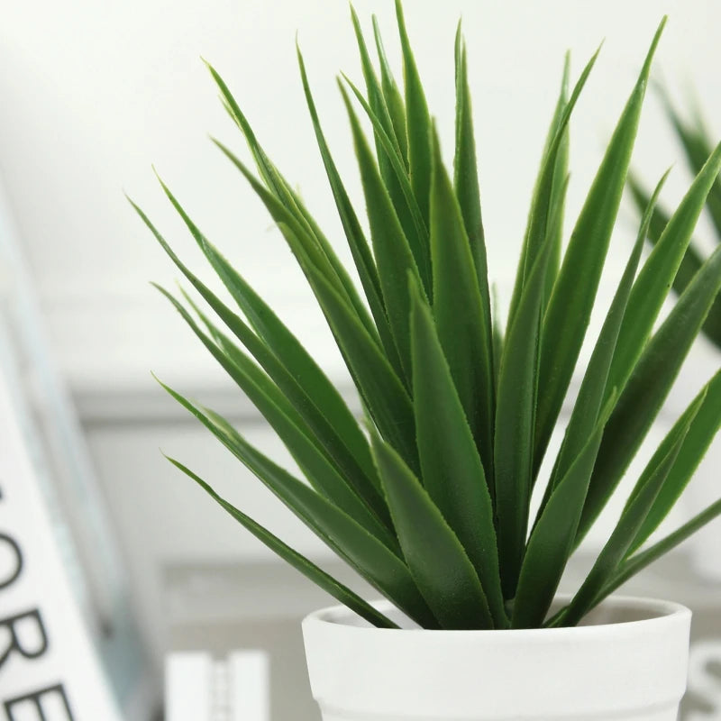 A close-up of a small, realistic artificial green grass plant in a simple, matte white ceramic pot.