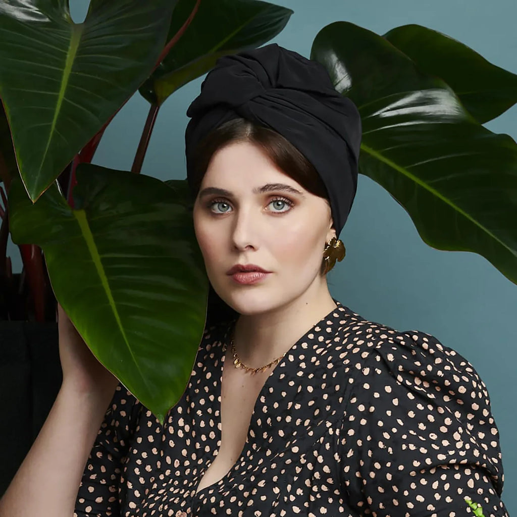A woman wears a sophisticated black vintage-style turban with a knotted front detail, set against large green leaves.