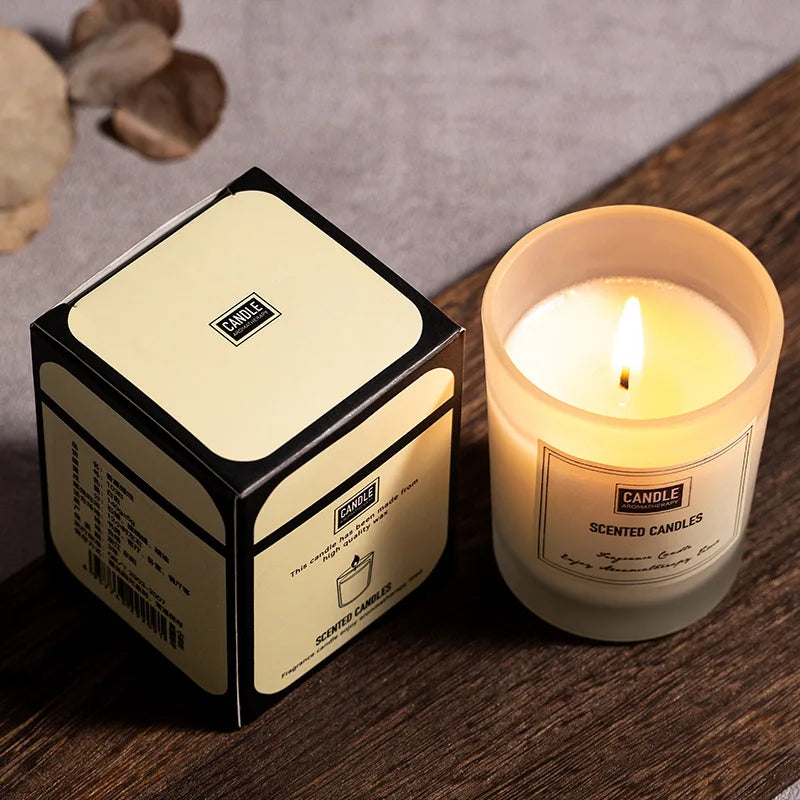 Lit scented candle in a frosted glass jar next to its cream and black presentation box on a wooden surface.